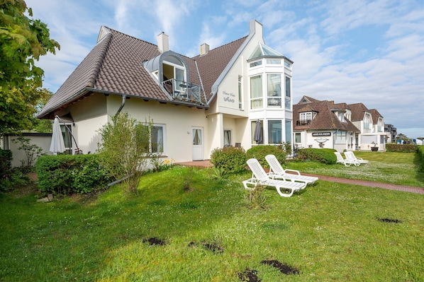 Une maison dotée d’une grande baie vitrée, d’un espace extérieur aménagé avec des chaises blanches, et d’une pelouse bien entretenue.