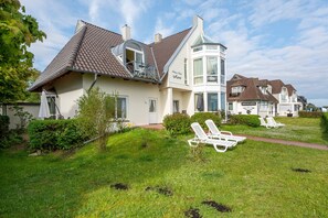 Une maison dotée d’une grande baie vitrée, d’un espace extérieur aménagé avec des chaises blanches, et d’une pelouse bien entretenue.