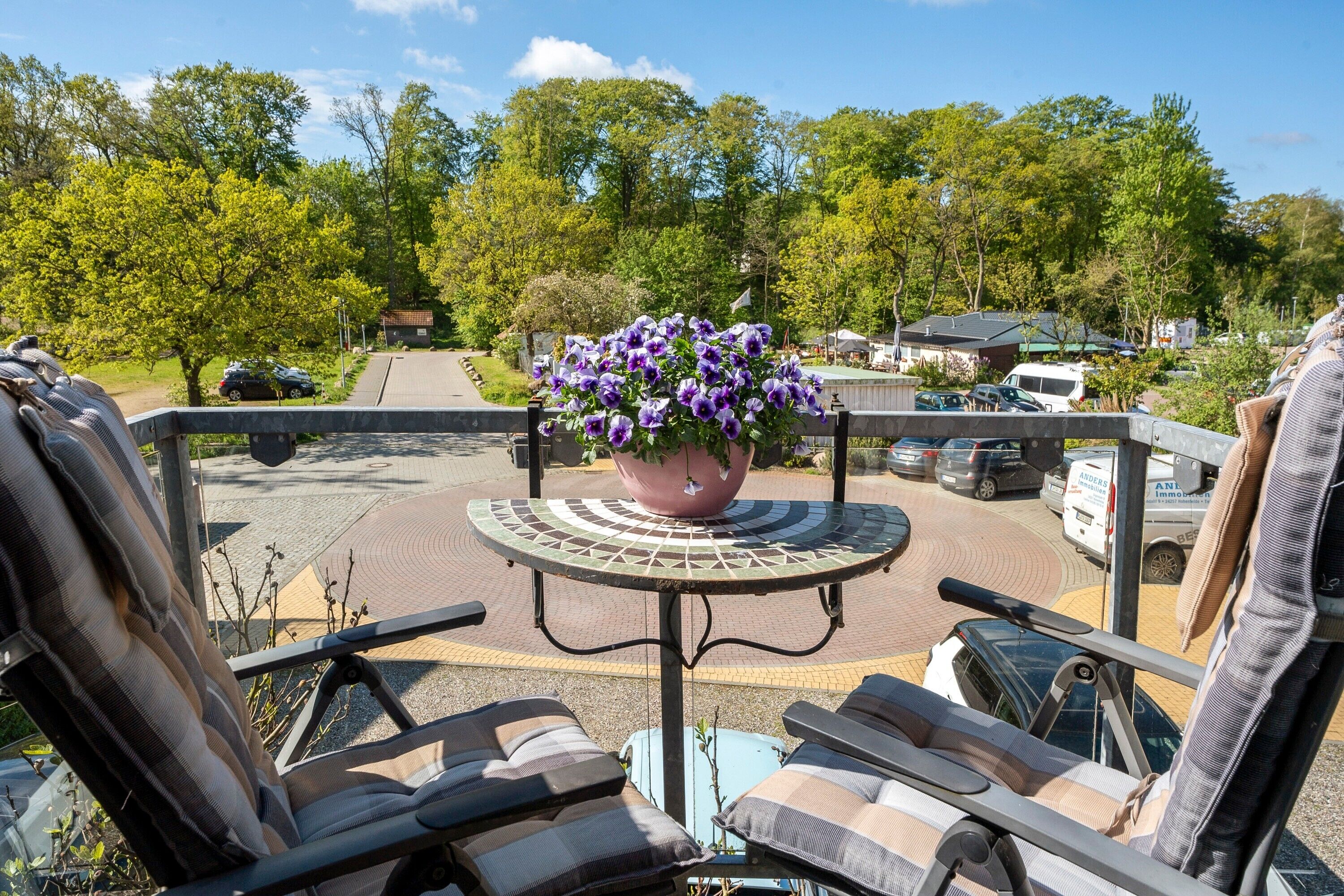 Ein Balkon mit zwei Liegestühlen, einem Topfpflanzen und Blick auf einen Parkplatz mit Bäumen und Gebäuden.