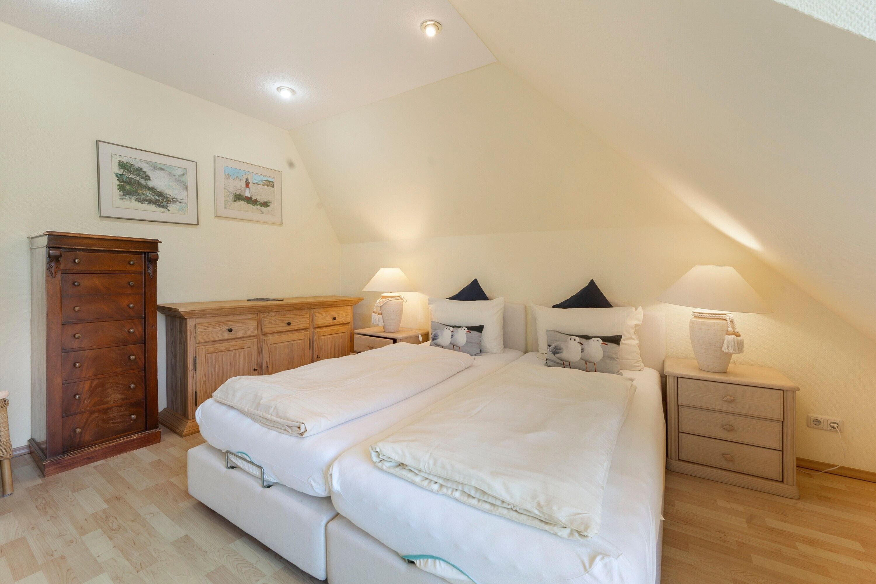 Une chambre à coucher avec un lit double, deux tables de chevet avec des lampes, une commode en bois et deux tableaux encadrés accrochés au mur.