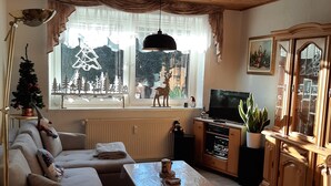 Una sala de estar con sofá, una mesa de centro, televisor, aparador y ventana con vista a árboles y una decoración de reno.