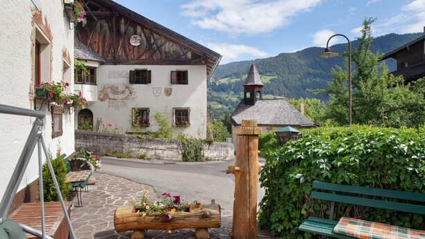 Un bâtiment traditionnel avec une façade en bois, un banc en pierre et un jardinier en forme de tronc d’arbre, également en bois, où sont plantées des fleurs.