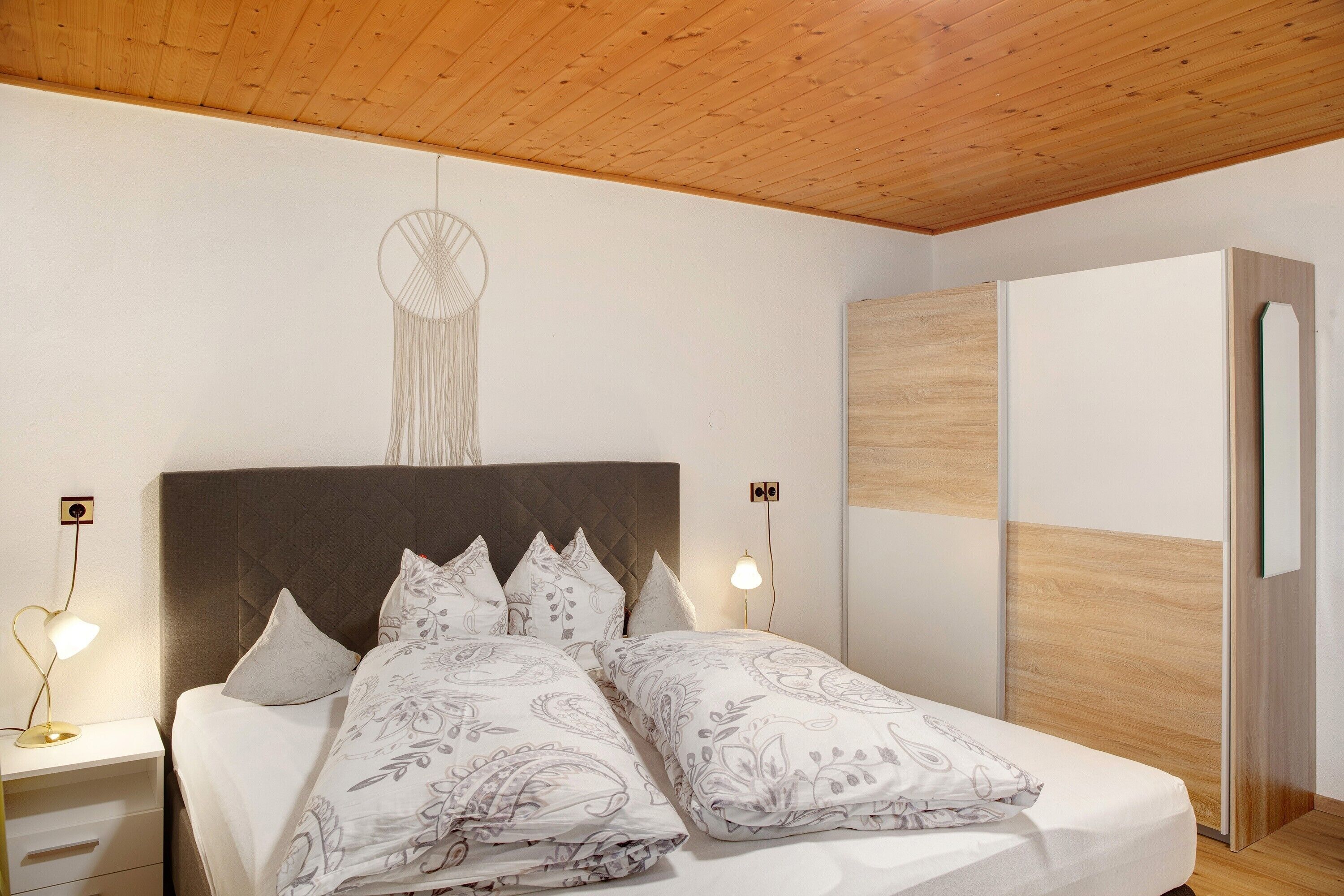 Quarto com cama, luminárias de cabeceira, guarda-roupa e um objeto decorativo na parede.