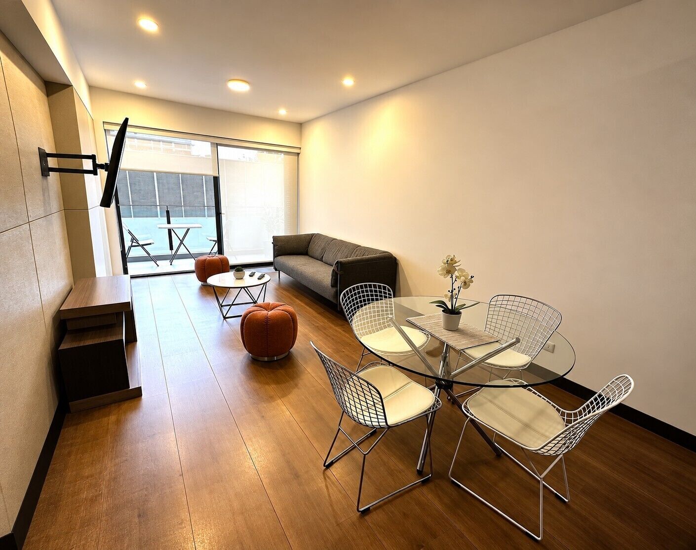 Un moderno salón con una mesa de comedor de vidrio, sillas de metal tipo alambre, un sofá y un pequeño otoman naranja.