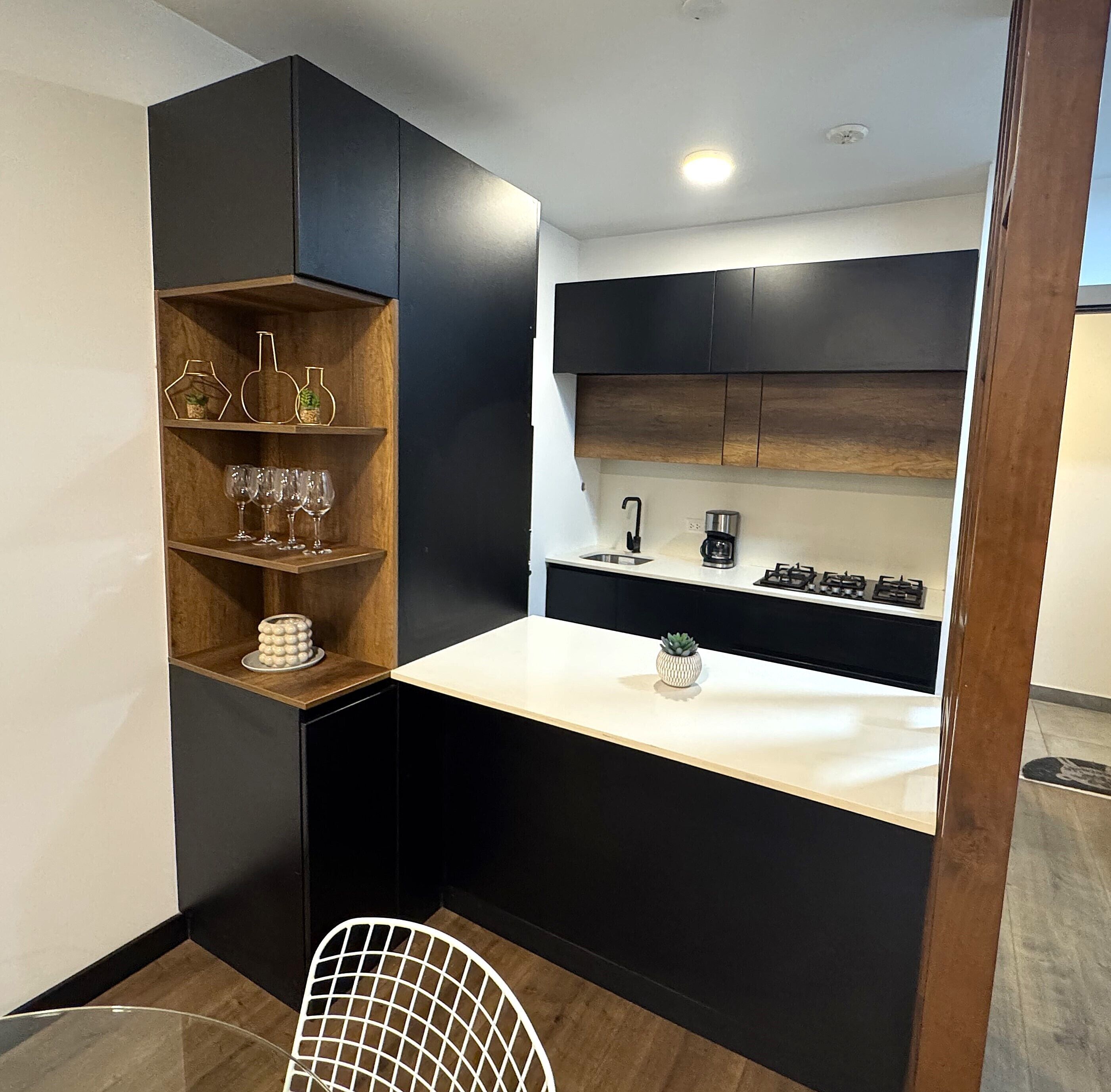 A compact kitchen with dark cabinetry, a white countertop, and a small shelf with decorative items.