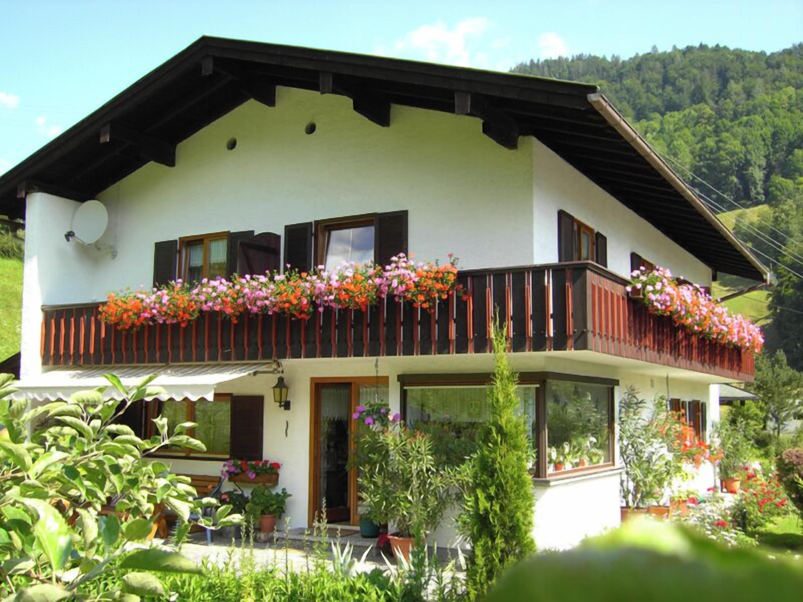 Une maison à deux étages dotée d’un balcon orné de fleurs, entourée d’une végétation luxuriante.