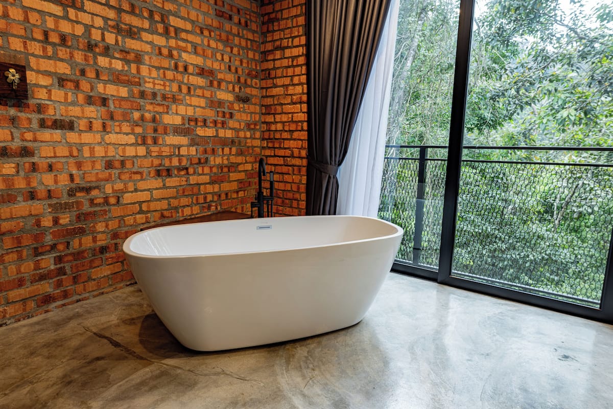 Un baño moderno con una bañera blanca grande, paredes de ladrillo y vistas a la vegetación a través de amplias ventanas.