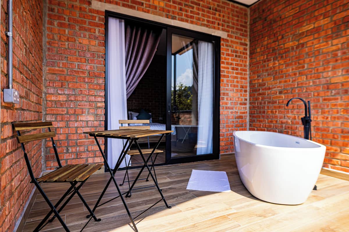 Un pequeño patio amurallado con pared de ladrillo, una mesa y sillas de madera, una bañera blanca grande y una puerta corrediza de vidrio con cortinas.