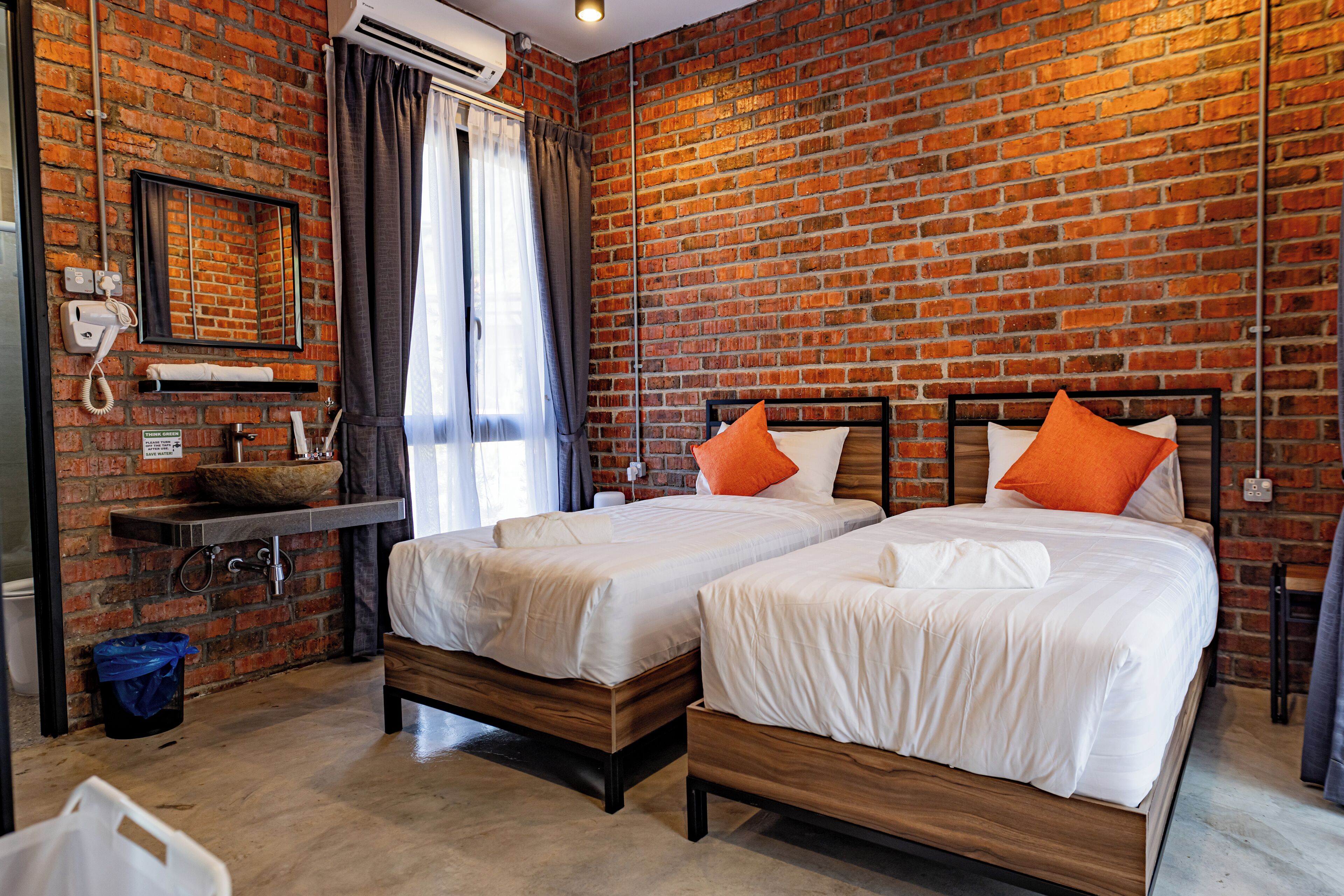 A hotel room with a brick wall, two beds with white bedding and orange pillows, a wooden bedside table, a mirror, and a window with curtains.