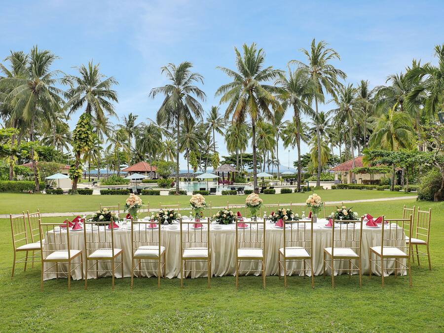 A formal outdoor dining setup with tables and chairs, surrounded by lush greenery and palm trees.