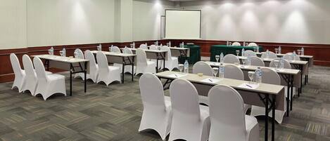 A conference room with multiple tables and chairs, equipped with water bottles and papers, and a projection screen.