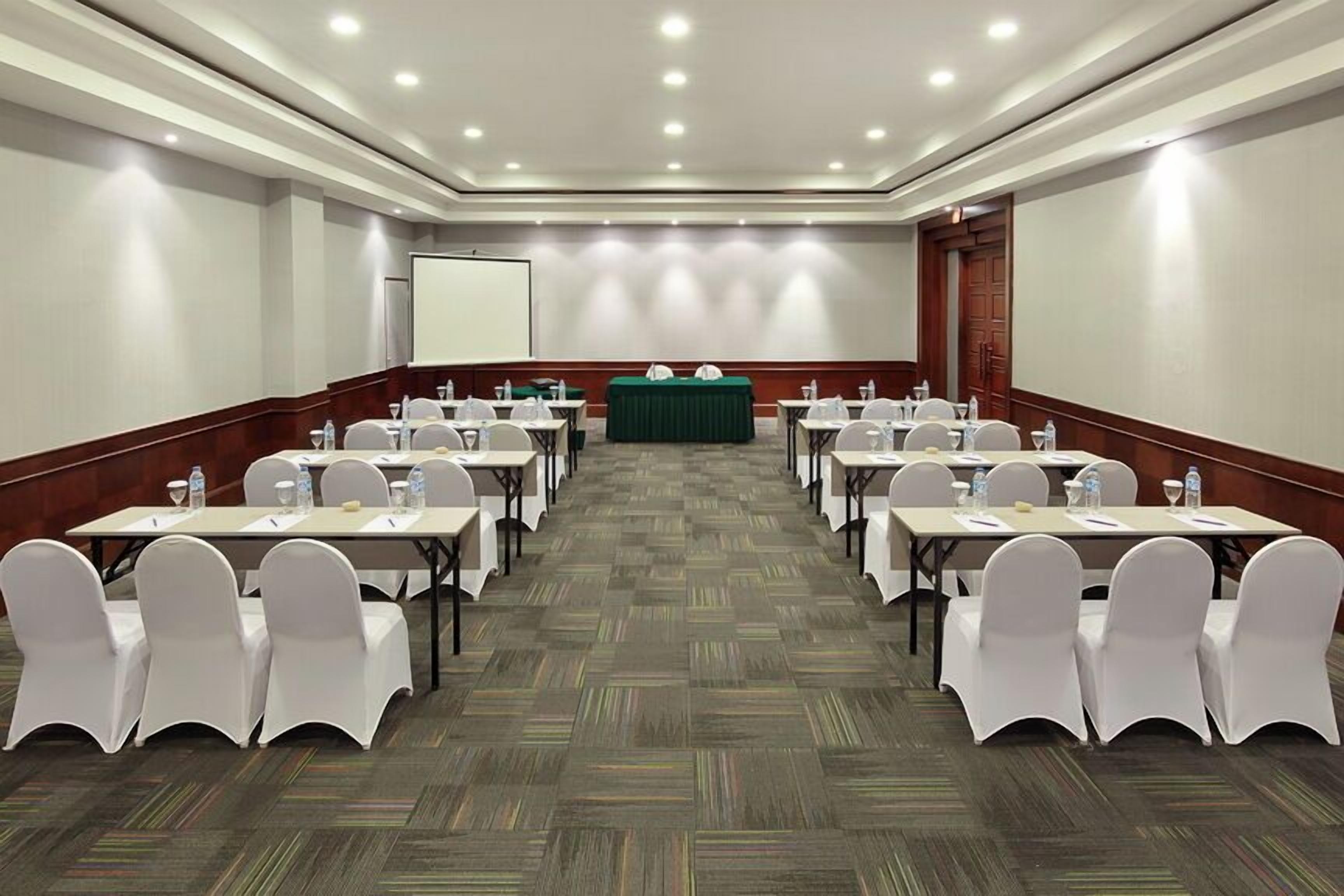 Una sala de conferencias con filas de mesas y sillas, una pantalla de proyector y una mesa verde al frente.