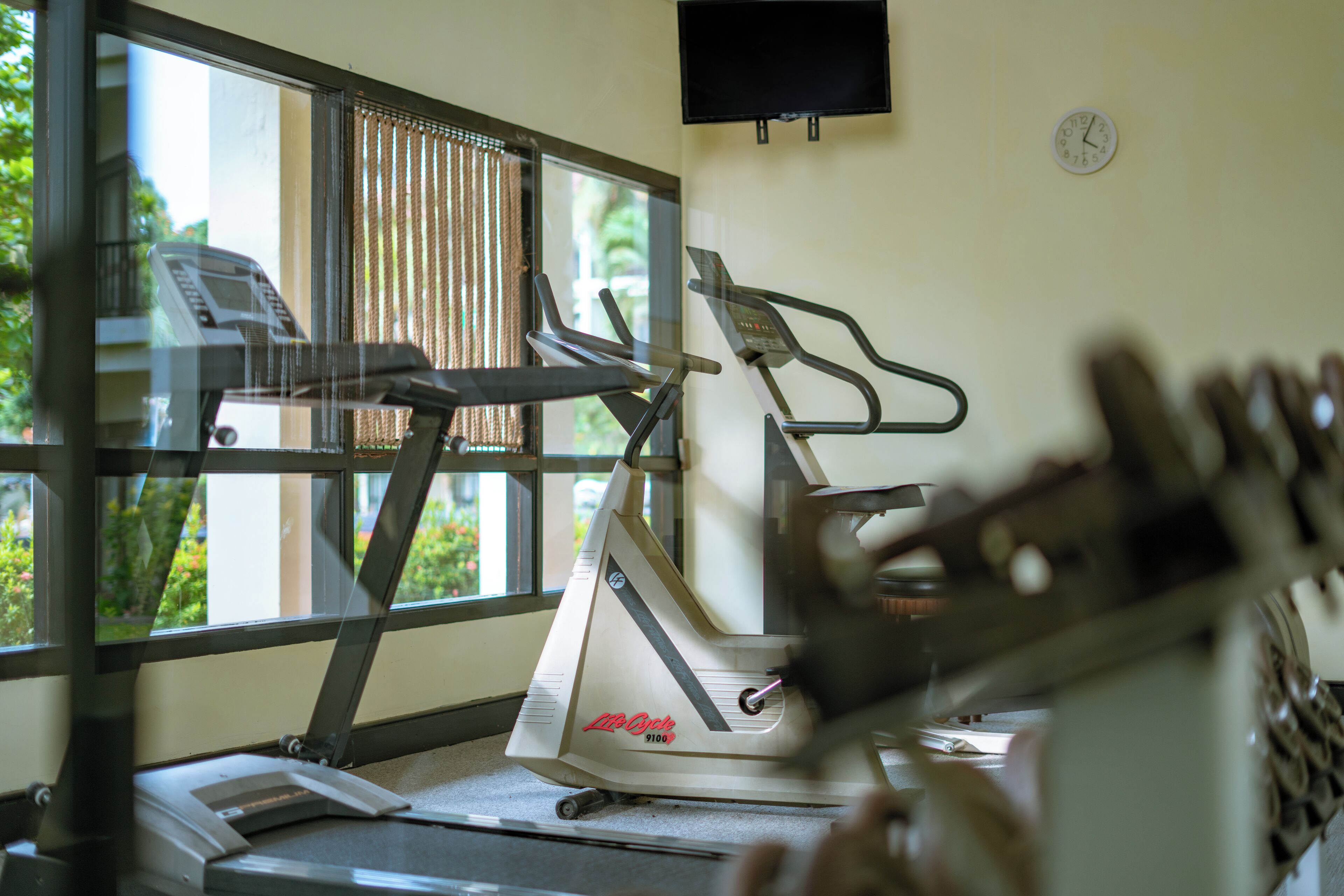 Una cinta de correr en una habitación con ventana, televisor de pared y un reloj.