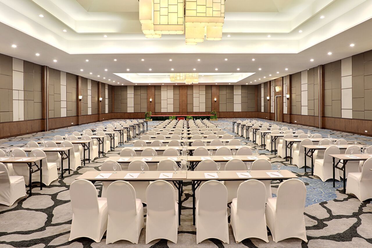 Un amplio salón de conferencias con filas de mesas y sillas, equipado con botellas de agua y documentos, y un escenario al frente.