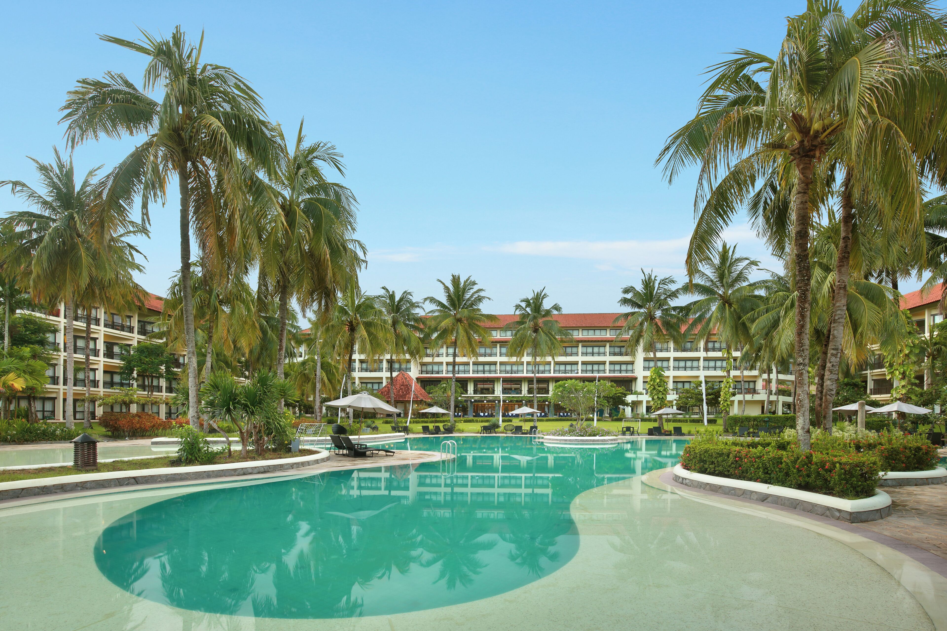 A resort pool with palm trees, a building with a red roof, and a clear blue sky.