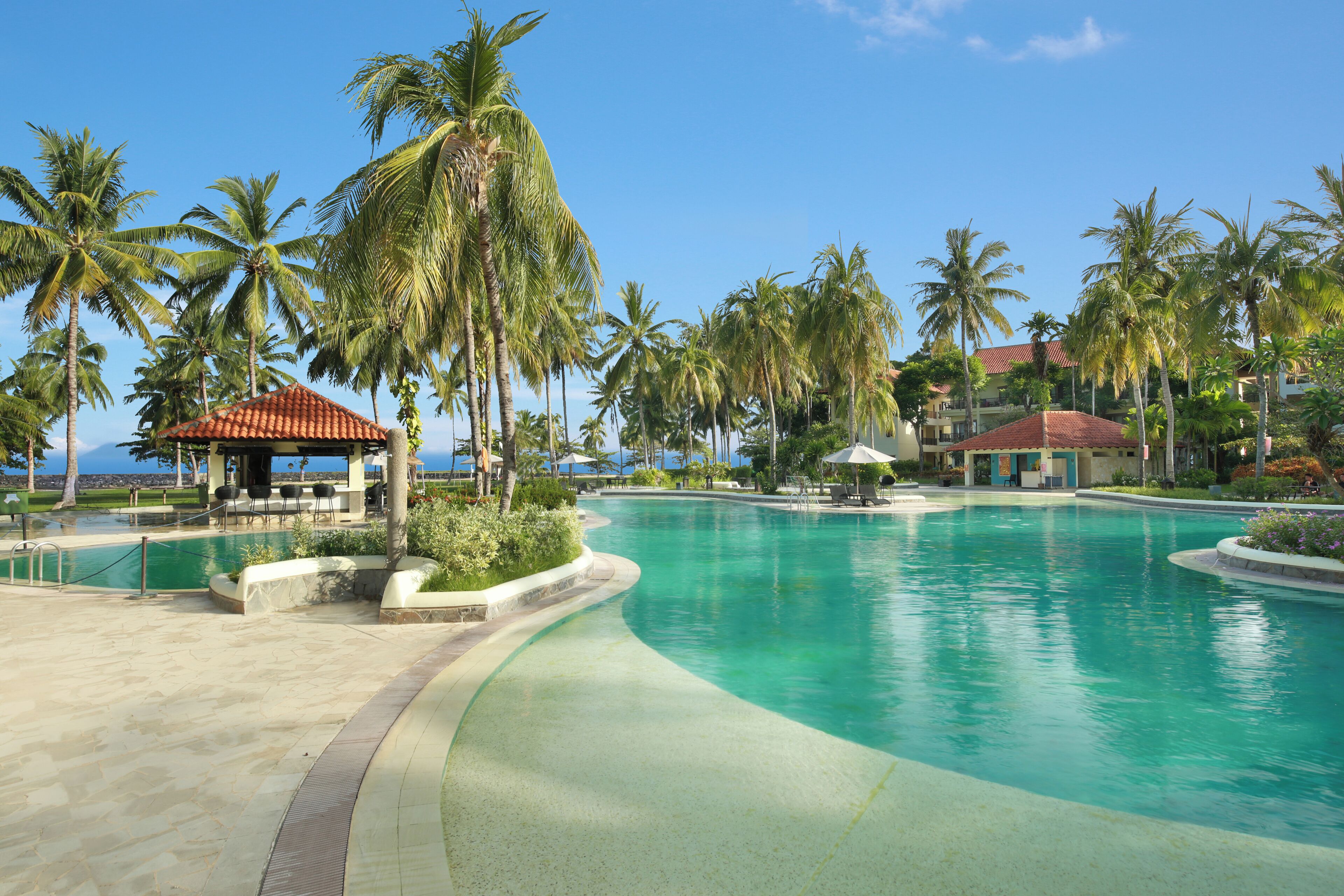 Una piscina di un resort con palme, una zona relax coperta e vista sull'oceano.