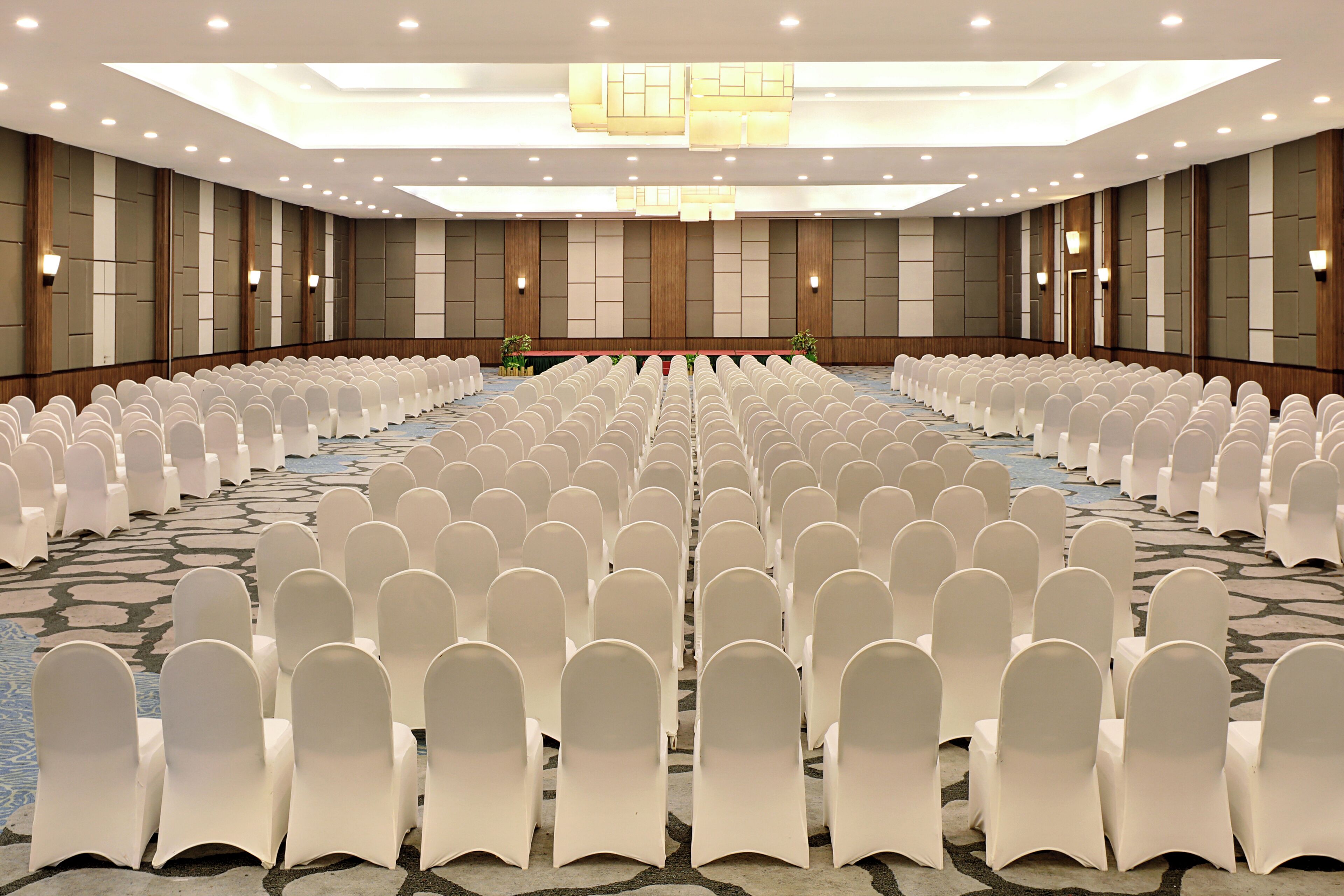 Un amplio salón de conferencias con filas de sillas blancas, una larga mesa y una alfombra estampada.