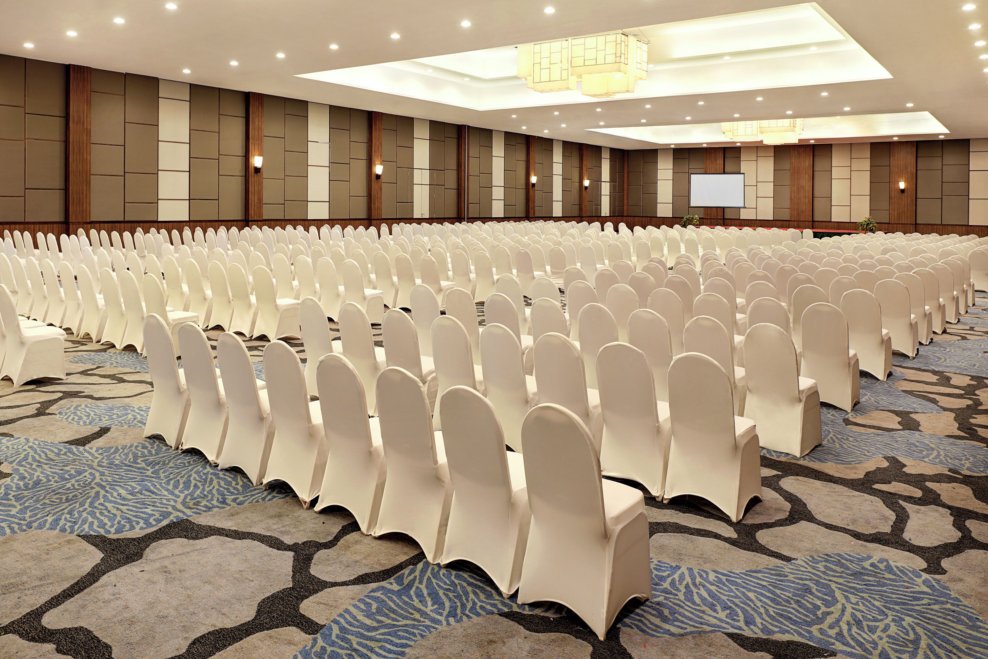 A large conference room with rows of white chairs, a projector screen, and a patterned carpet.