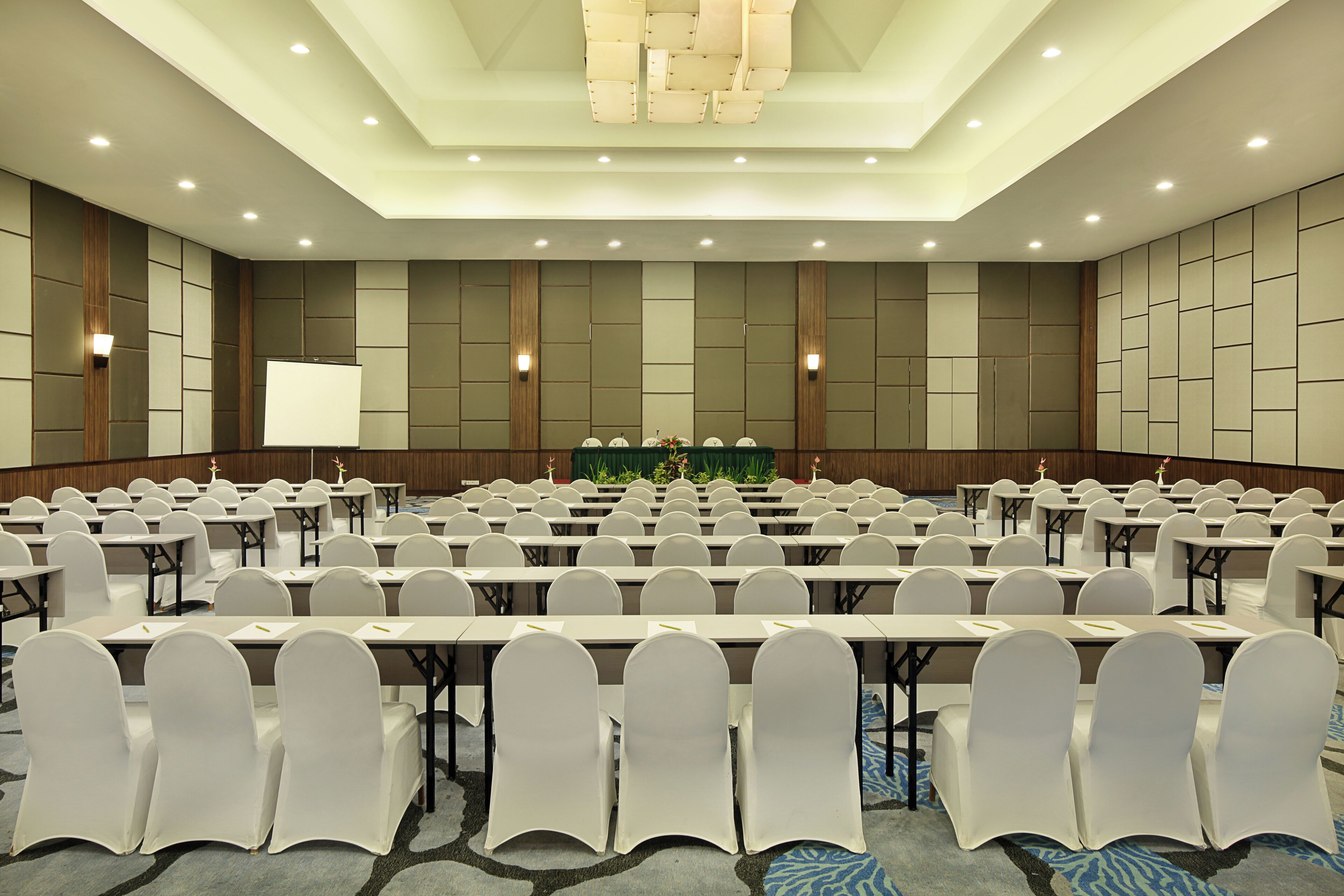 Un salón de conferencias con filas de mesas y sillas, un atril y una pantalla de proyector.