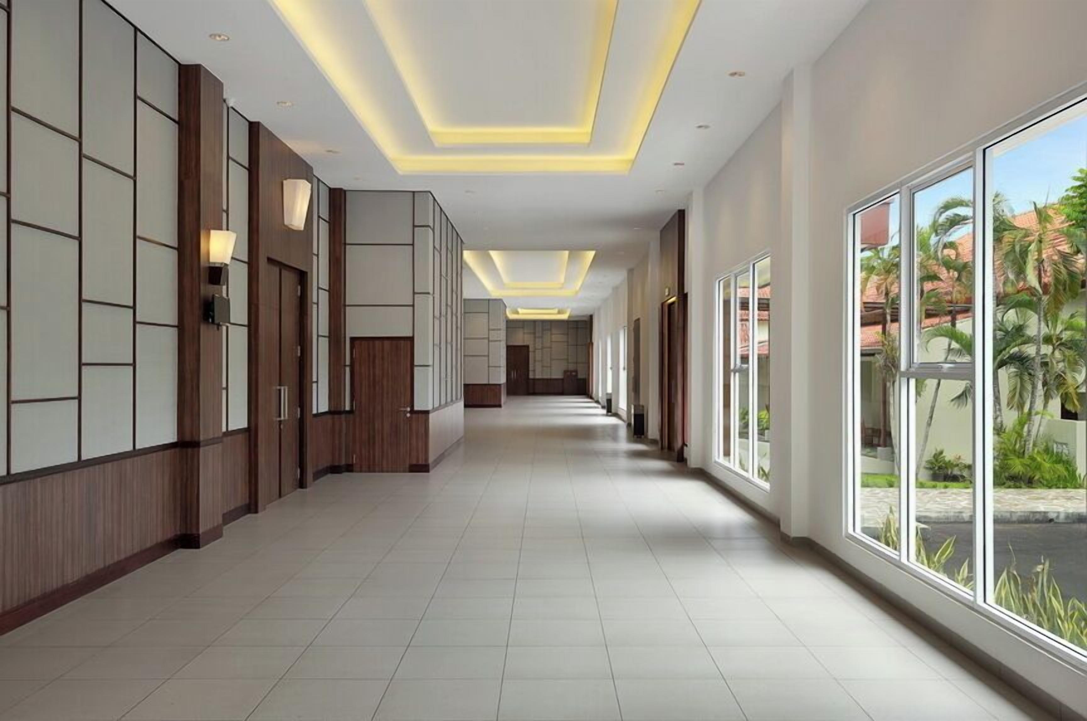 A modern hotel corridor with wooden paneling, recessed lighting, and large windows offering views of palm trees.
