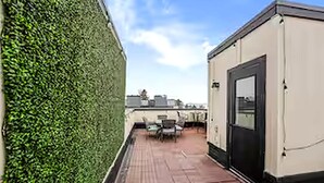 A rooftop patio with a table and chairs, a door, and a green hedge. - West Bay Hideaway | Rooftop Condo Near Beach/DT TC (Traverse City)