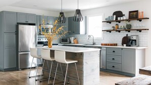 A modern kitchen with grey cabinets, a stainless steel refrigerator, a central island with bar stools, pendant lighting, and a wooden coffee table. - Woodland Chase - Pool + Hot Tub by Wineries (Prince Edward)