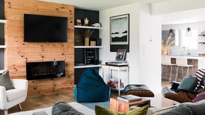 A modern living room with a flat-screen TV, wooden wall paneling, a fireplace, and a kitchen area visible through an open doorway. - Woodland Chase - Pool + Hot Tub by Wineries (Prince Edward)