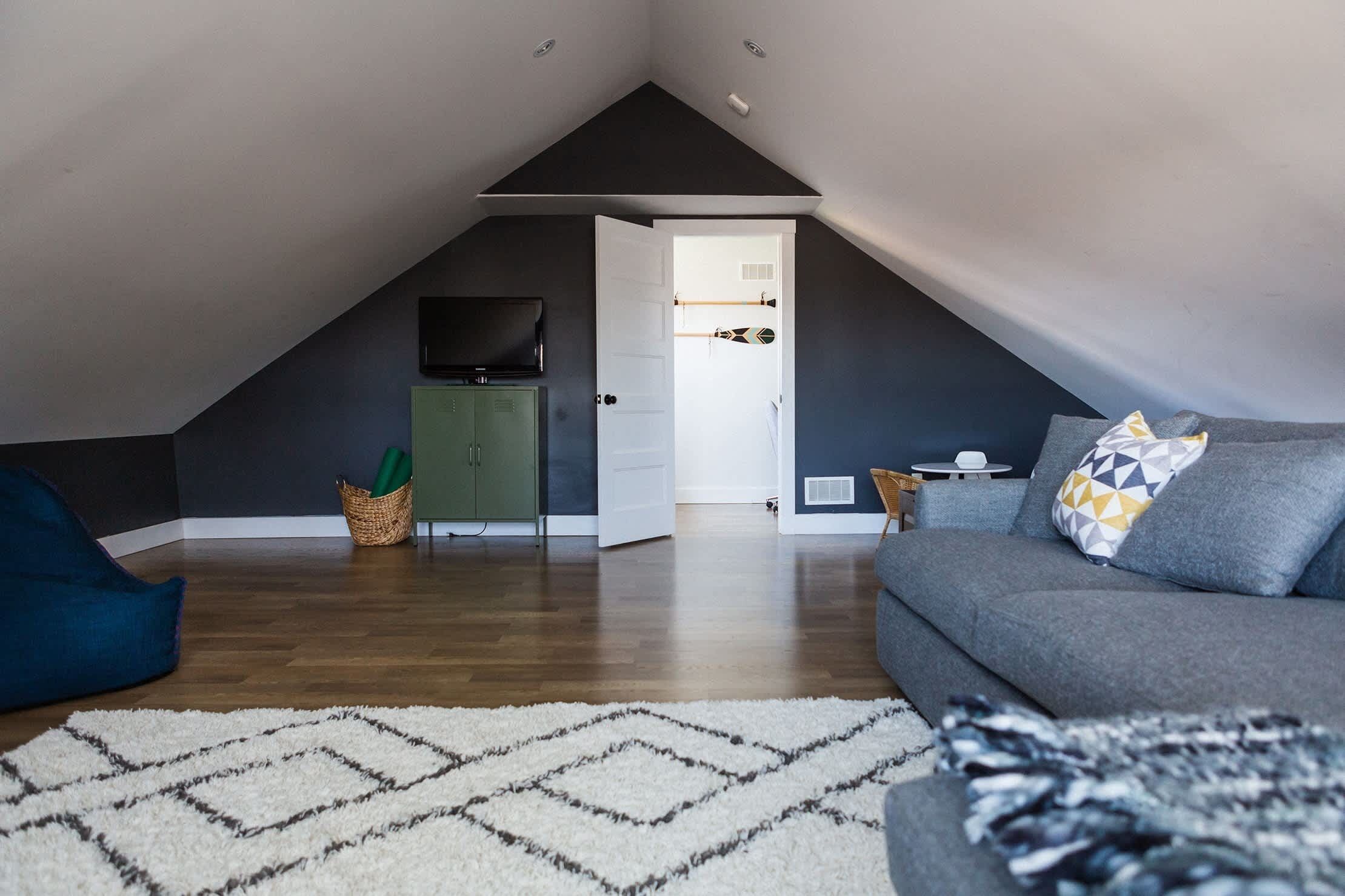 Ein Dachzimmer mit schrägen Wänden, einem grauen Sofa, einem weißen Teppich, einem grünen Schrank und einem Fernseher.