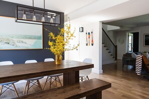 A dining area with a wooden table, chairs, a vase with yellow flowers, a painting of a beach, and a staircase leading to another room. - Woodland Chase - Pool + Hot Tub by Wineries (Prince Edward)
