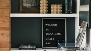 A room with a dark green wall, wooden paneling, a white shelf with books, a framed picture, a welcome sign, and a small table with a record player. - Woodland Chase - Pool + Hot Tub by Wineries (Prince Edward)