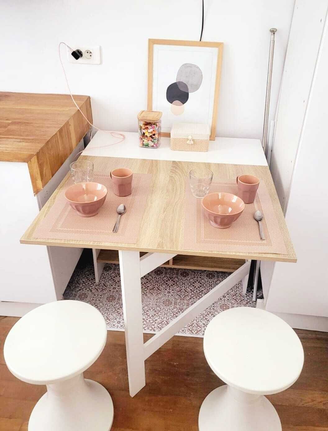A small dining table with pink placemats, two white stools, and a framed picture on the wall.