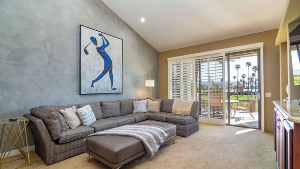 A spacious living room with a sectional sofa, a large painting, and a view of a golf course through the sliding glass doors. - Country Club Experience (Palm Desert)
