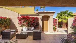An outdoor patio area with wicker furniture, a covered seating area, and vibrant flowering plants. - Country Club Experience (Palm Desert)