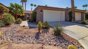 A residential house with a garage, surrounded by a landscaped garden with various plants and a rock pathway. - Country Club Experience (Palm Desert)