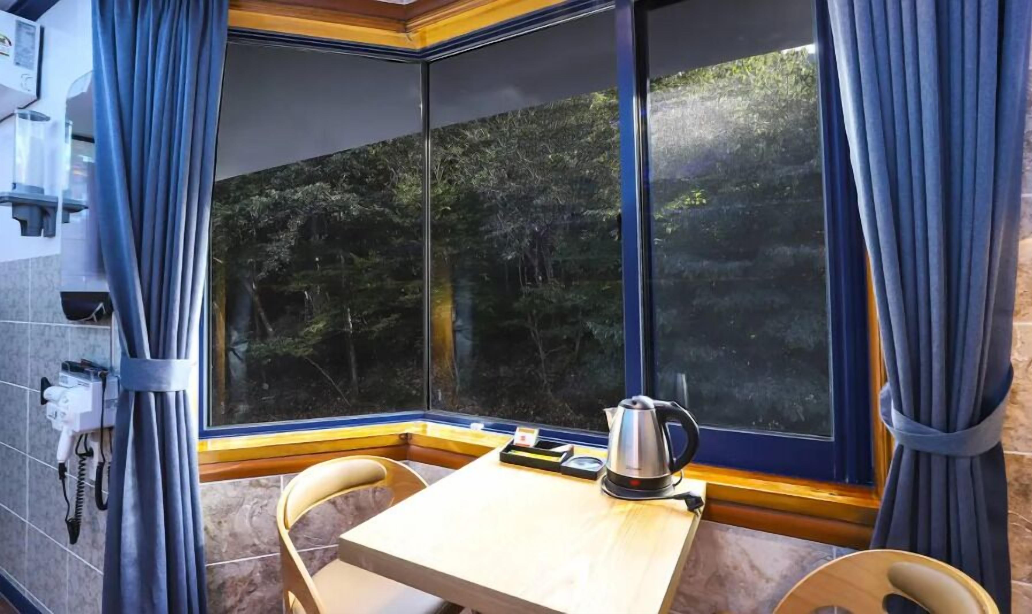 A corner room with a table, chairs, a kettle, and a view of a forest through large windows.