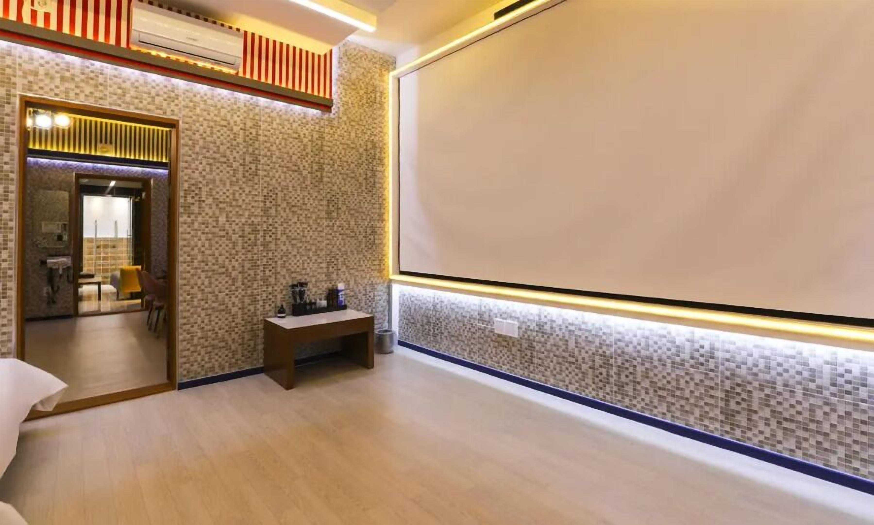 A hotel room with a large screen, a small table with a chair, and a bathroom visible through a glass door.