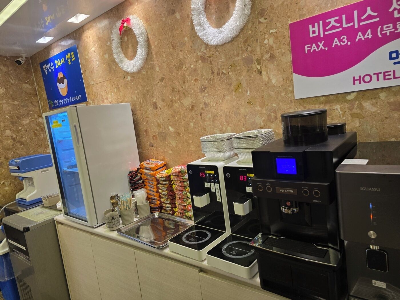 Zona de recepción de un hotel con un refrigerador, paquetes de bocadillos apilados, una máquina de café y un fax.