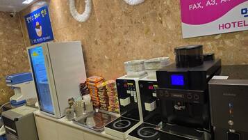 Zona de recepción de un hotel con un refrigerador, paquetes de bocadillos apilados, una máquina de café y un fax.