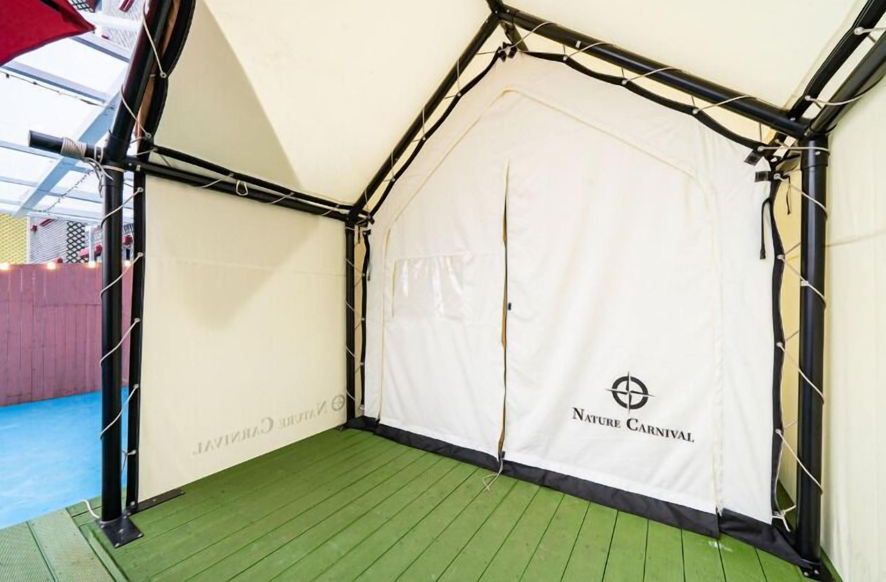 Una carpa con el texto "Nature Carnival" en el lateral, montada sobre un suelo verde con una pared blanca al fondo.