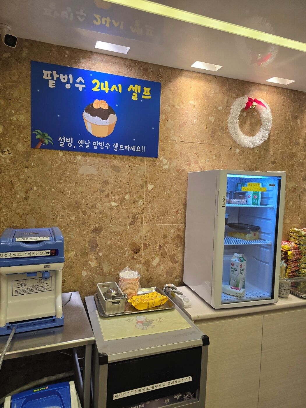 A hotel reception area with a refrigerator stocked with beverages, a blue machine with a yellow label, and a signboard advertising a 2-night stay promotion.