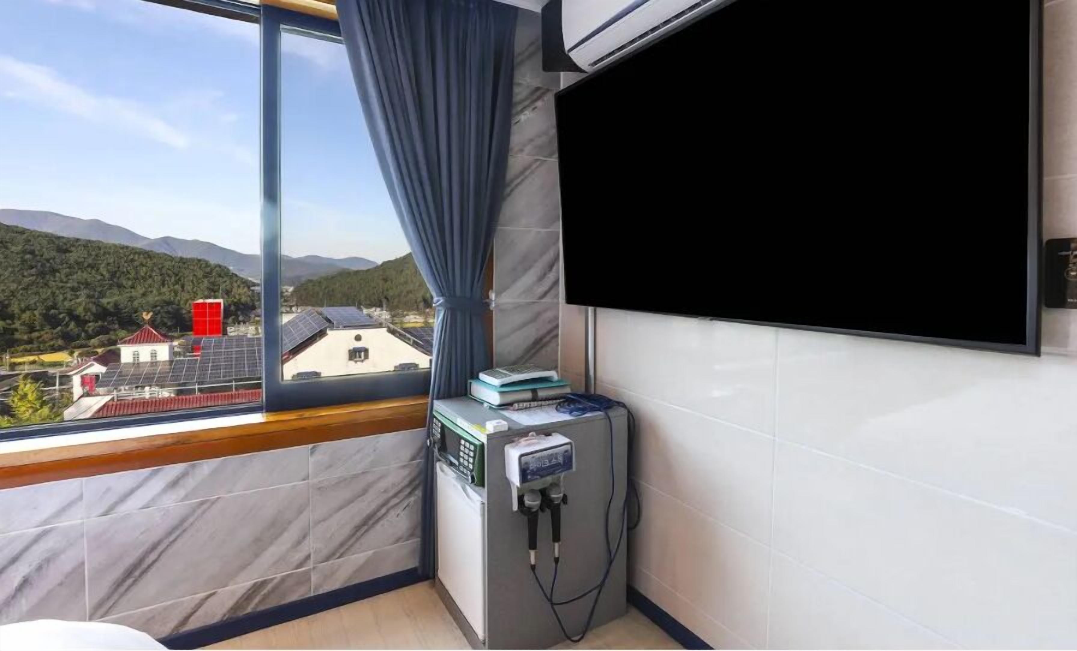 A hotel room with a large window offering a view of solar panels and buildings, a flat-screen TV, and a small table with books and a power strip.