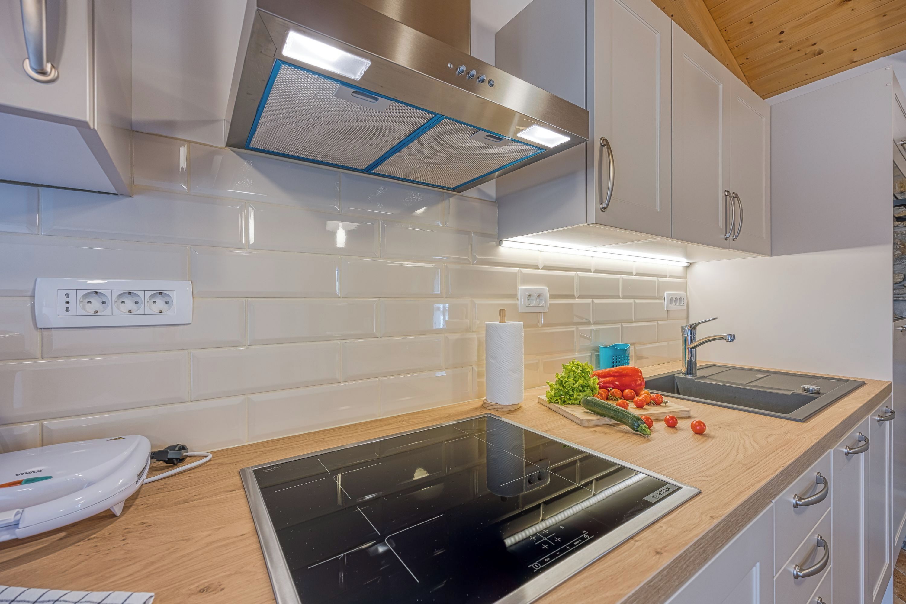 A modern kitchen with a black induction cooktop, stainless steel range hood, white cabinets, and a wooden countertop.