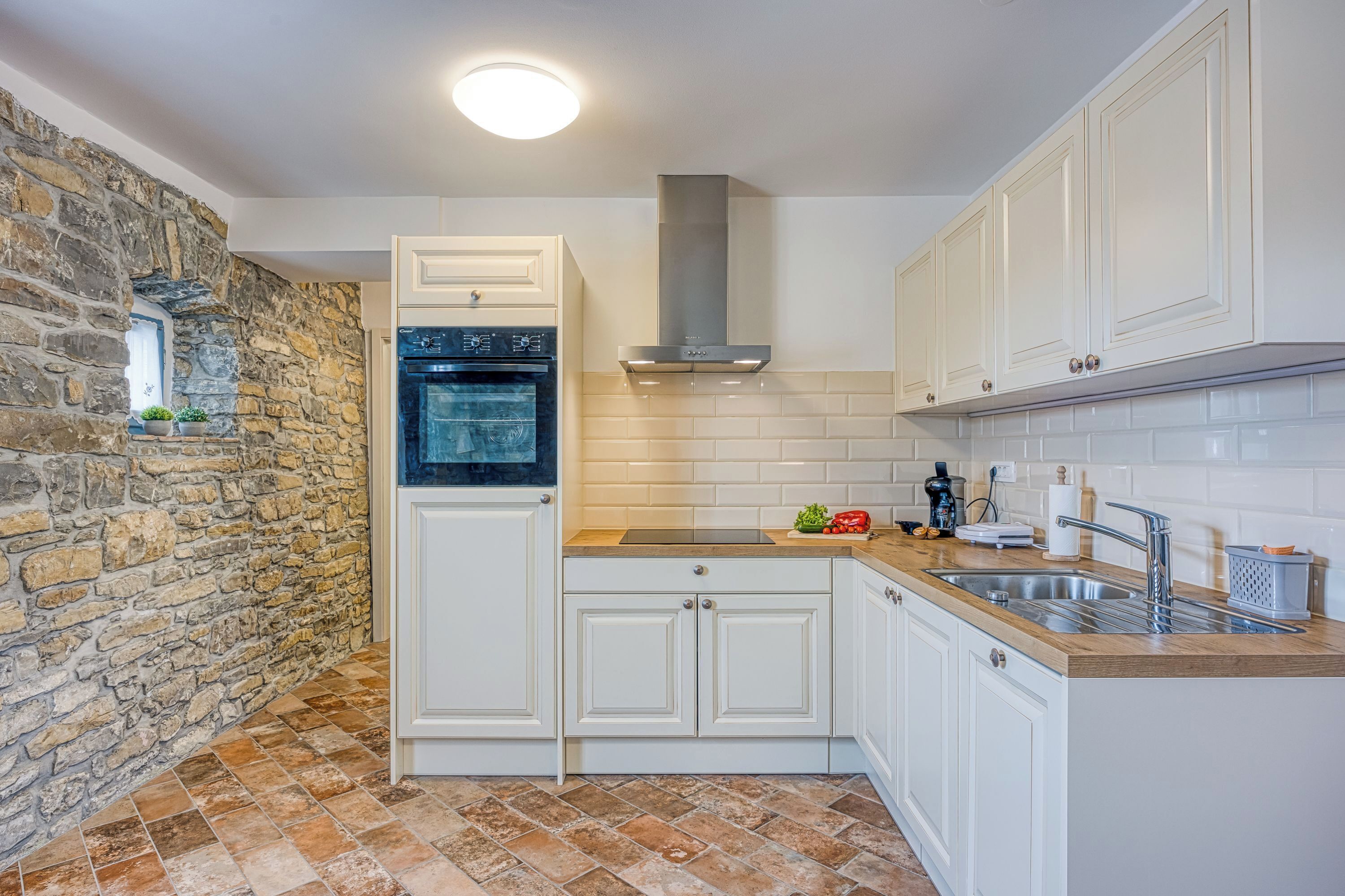 A kitchen with white cabinets, a stone wall, a built-in oven, a stainless steel range hood, and a wooden countertop.