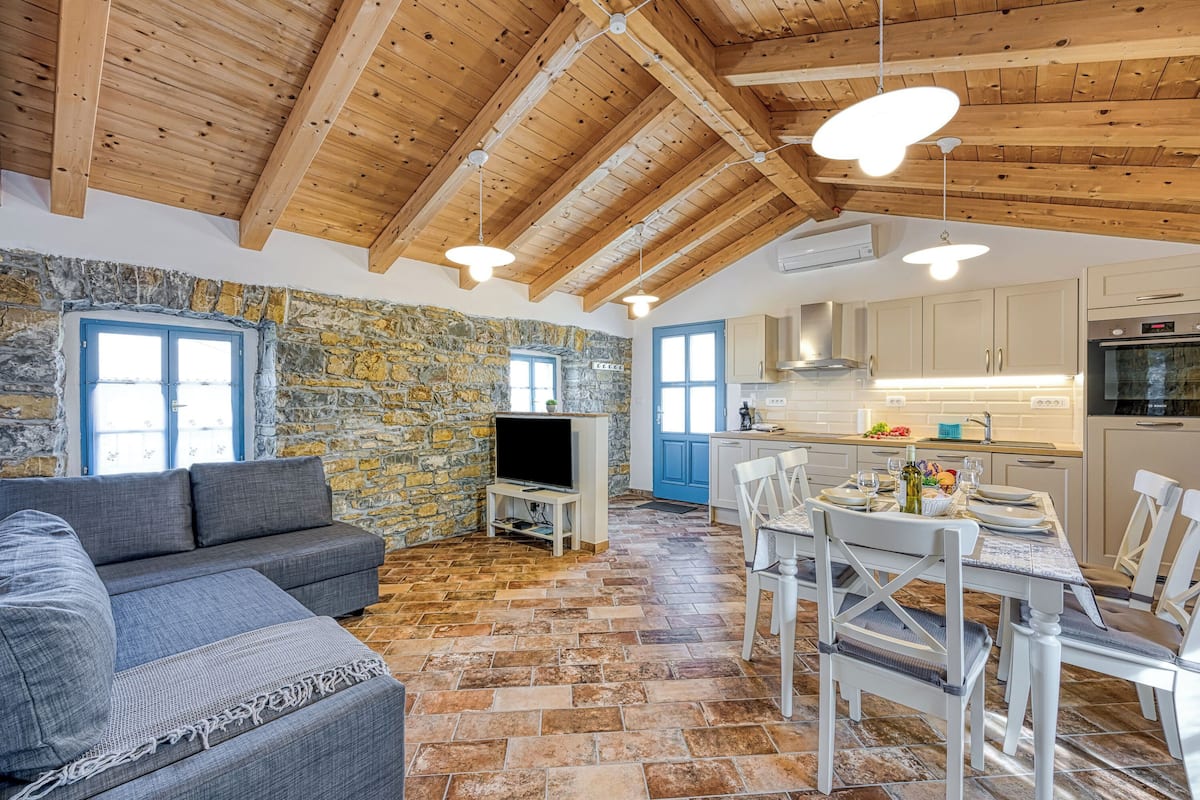 A cozy living area with a stone wall, wooden ceiling beams, a gray sofa, and a dining table with chairs.