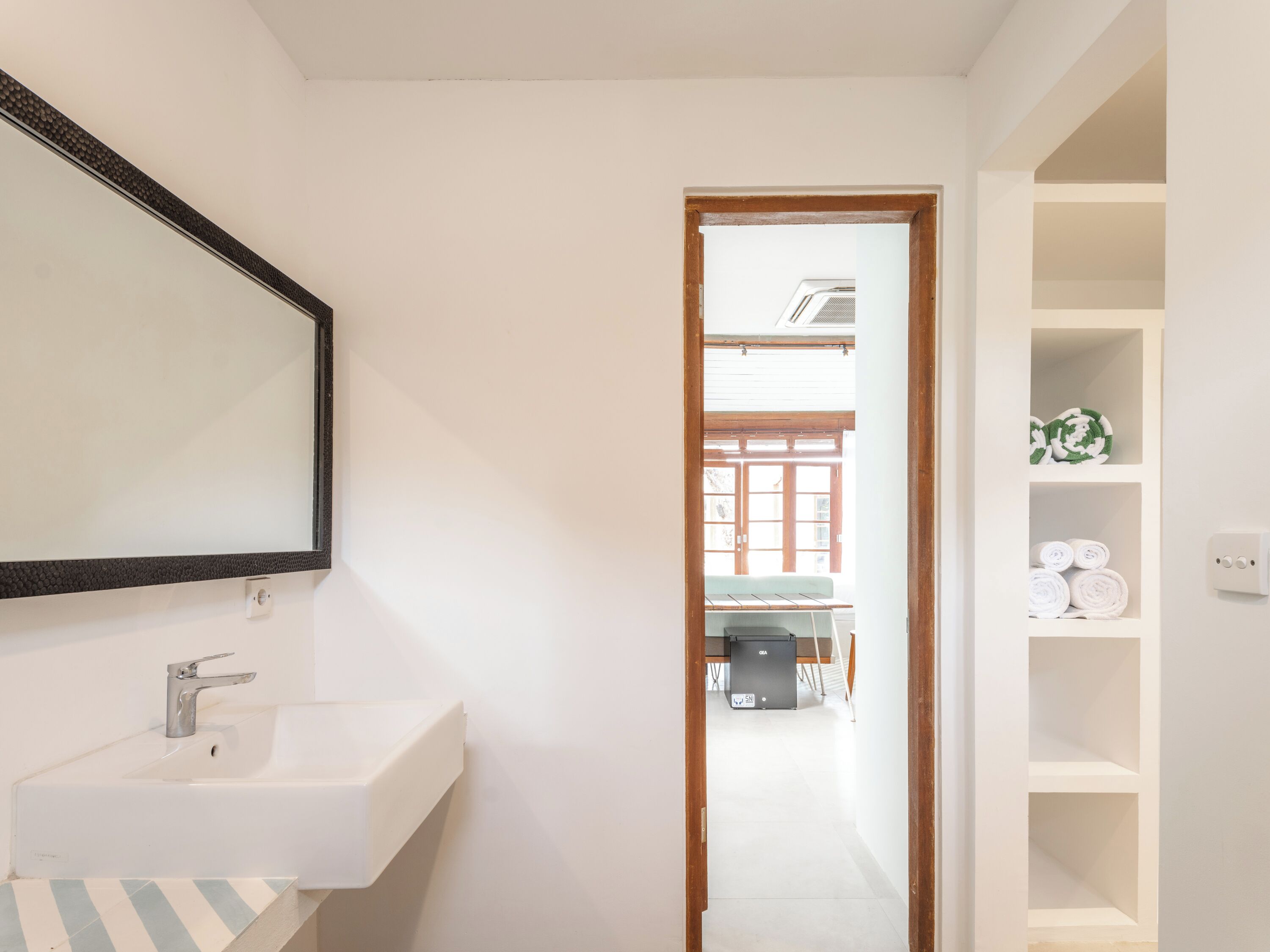 Une salle de bain avec un lavabo blanc, un miroir et une porte en bois donnant sur une pièce où se trouvent un lit et une fenêtre.