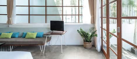 A room with a large window, a sofa with cushions, a TV on a stand, and a potted plant.