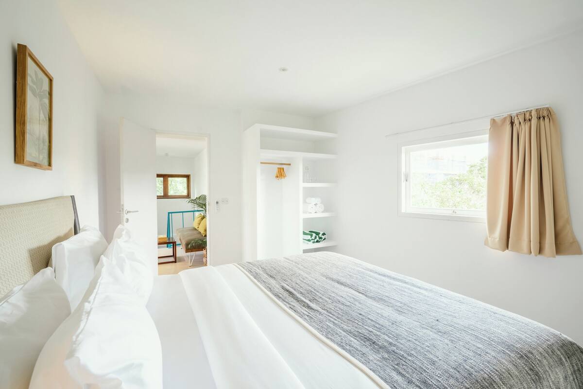 Een slaapkamer met een groot bed, een kledingkast, een raam met gordijnen en een deur naar een andere kamer.
