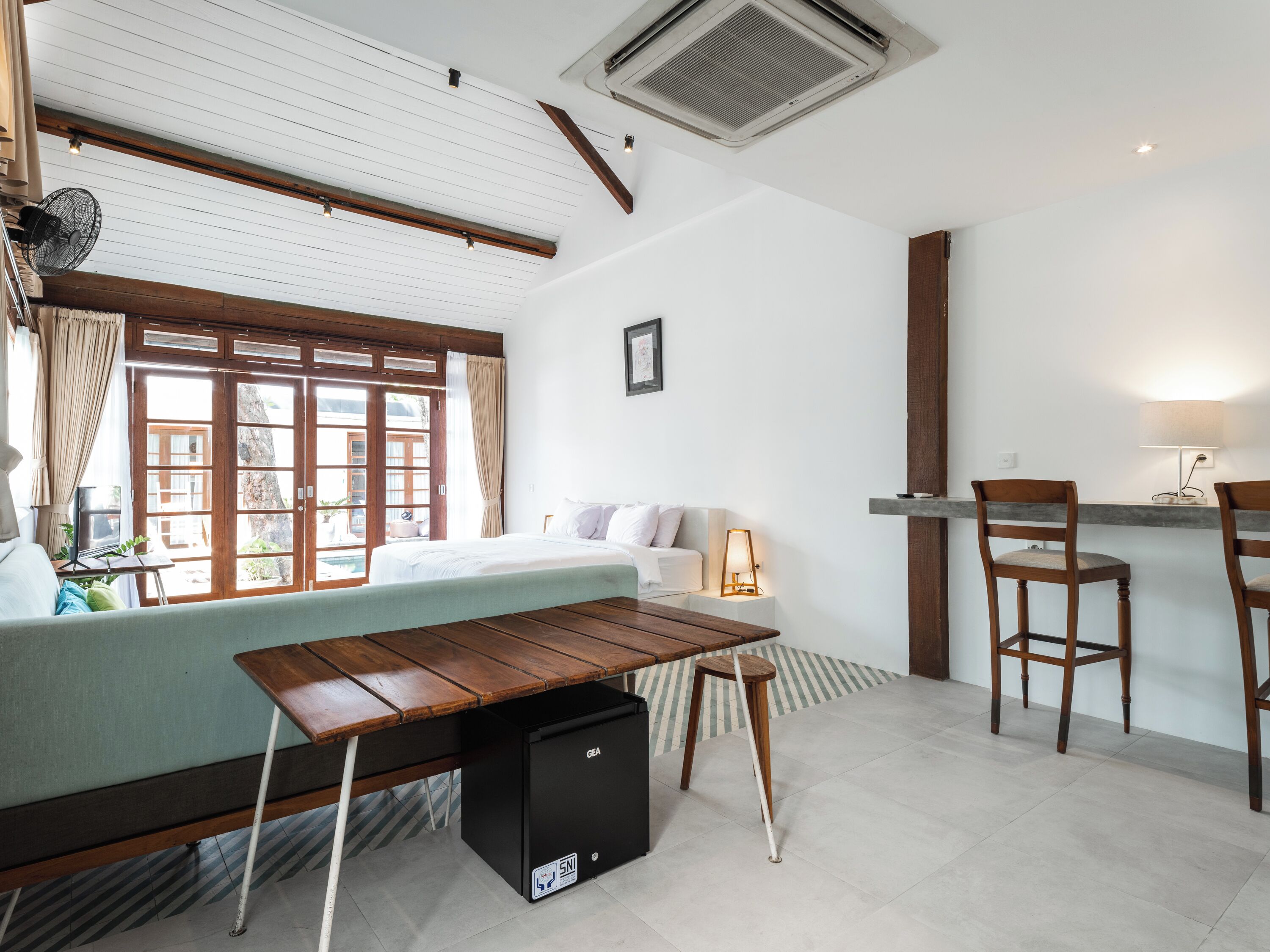 Een moderne, goed verlichte kamer met een bed, een bureau en een stoel. Er is een raam met gordijnen, een wandairconditioner en een klein tafeltje met een lamp.