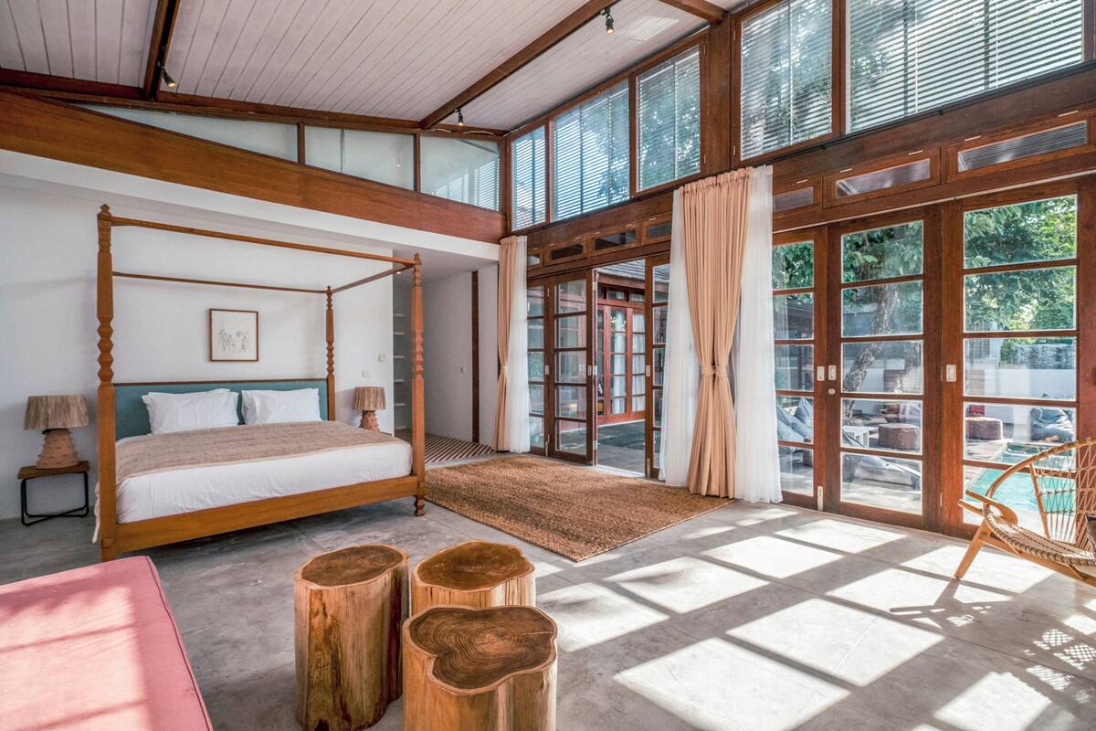 Een ruime slaapkamer met een hemelbed, houten nachtkastjes en een groot raam met uitzicht op de omgeving.