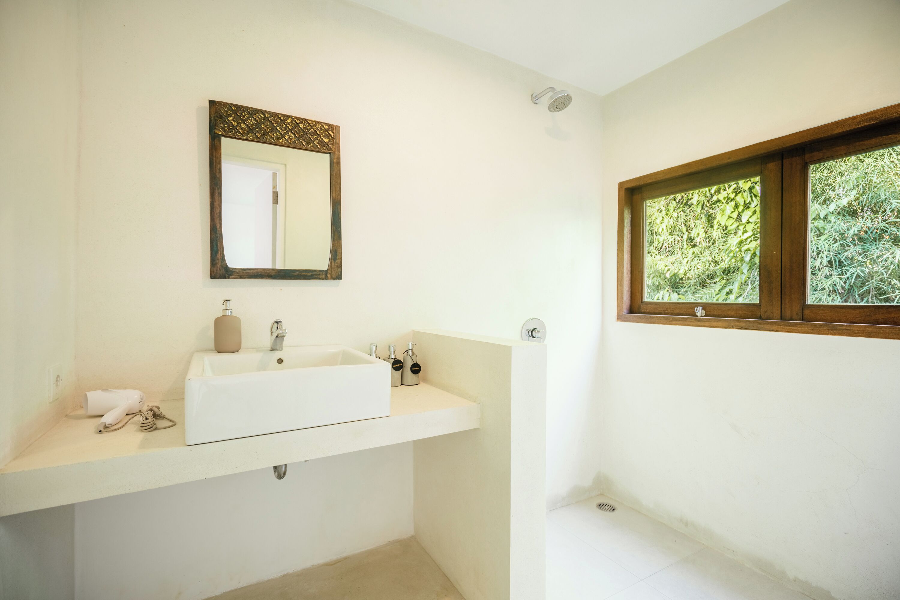 Un baño con un lavamanos blanco, un espejo con un marco decorativo, una ventana con vista a la vegetación y una zona de ducha.